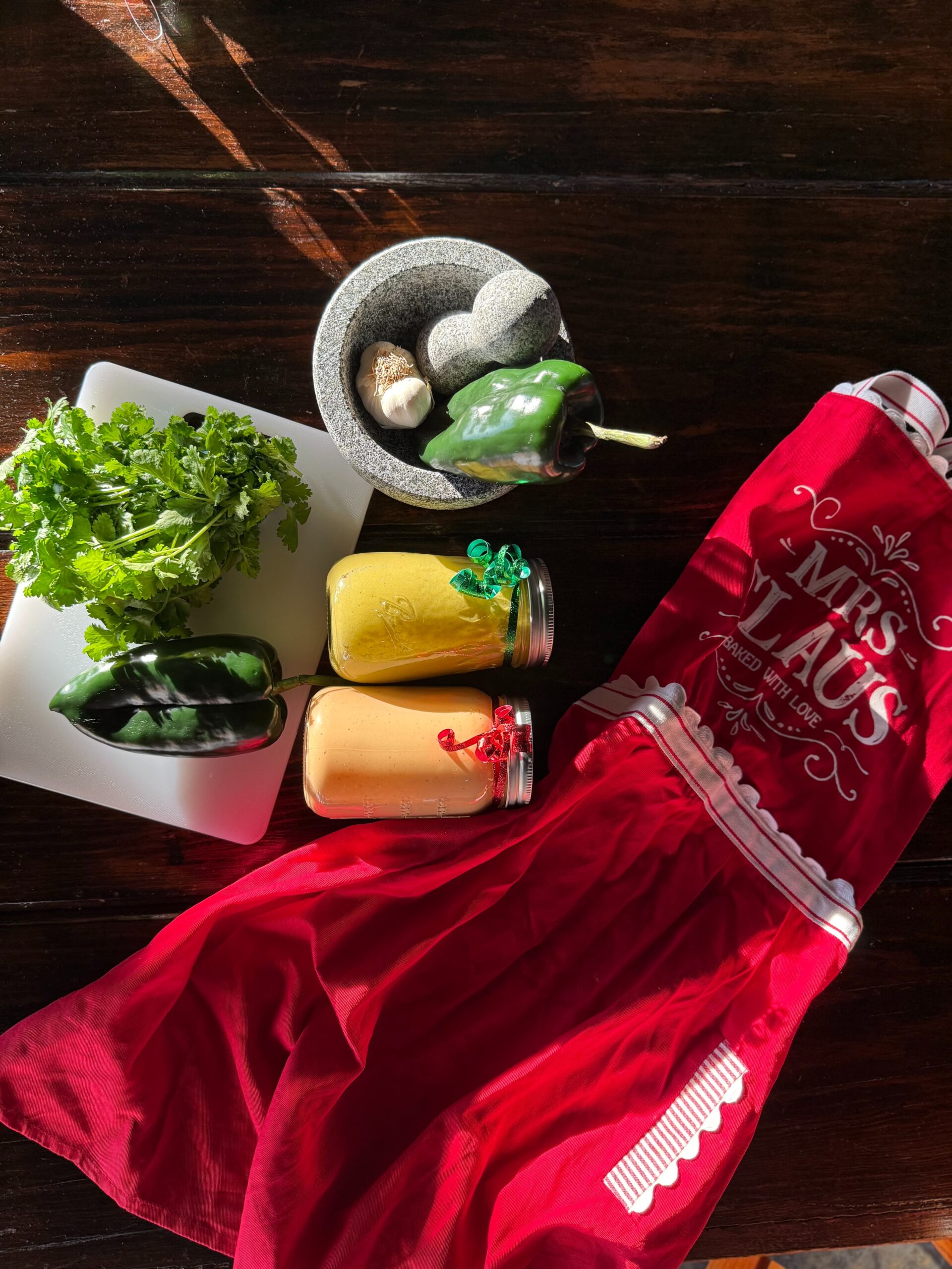Todo Natural chipotle cream and poblano cream jars with fresh poblano peppers, garlic, cilantro in molcajete, and Mrs. Claus Christmas apron on rustic wood table - Brownsville Texas holiday catering